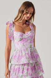 A woman is wearing a sleeveless dress with shoulder ties. The dress features a light, semi-sheer fabric with a floral print in shades of pink, purple, and green. It has a fitted bodice with a sweetheart neckline decorated with a delicate scalloped trim, and a tiered, ruffled skirt that adds volume and movement.